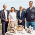 Vier Personen stehen hinter einem Tisch mit verschiedenen Gläsern und Körben. Im Hintergrund sind eine Wiese und Hügel zu sehen. Alle tragen formelle Kleidung.