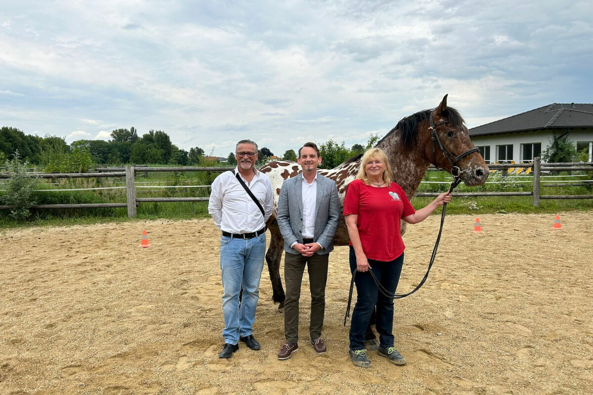 Drei Personen stehen auf einem Sandplatz vor einem Pferd. Eine Person hält das Pferd am Zügel. Im Hintergrund ist ein Holzzaun und ein Gebäude zu sehen. Bewölkter Himmel.