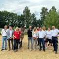 Eine Gruppe von Menschen steht draußen auf einem sandigen Platz, neben einem großen, braunen Pferd. Im Hintergrund sind Bäume und ein bewölkter Himmel zu sehen. Die Personen lächeln in die Kamera.