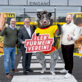 Drei Männer stehen neben einem Wildschwein-Maskottchen in gelbem Trikot vor einem Supermarkt-Eingang. Einer der Männer hält ein Schild mit der Aufschrift „I leb’ für mein’ Verein!“ Alle lächeln und zeigen mit Daumen hoch.