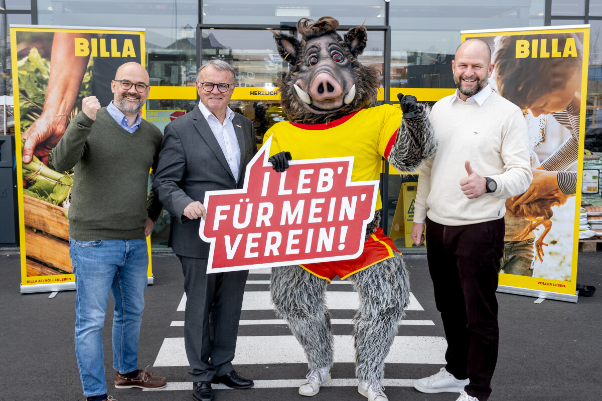 Drei Männer stehen neben einem Wildschwein-Maskottchen in gelbem Trikot vor einem Supermarkt-Eingang. Einer der Männer hält ein Schild mit der Aufschrift „I leb’ für mein’ Verein!“ Alle lächeln und zeigen mit Daumen hoch.