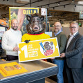 Drei Männer stehen in einem Supermarkt neben einer Person im Wildschweinkostüm. Sie halten ein großes gelbes Plakat mit dem Text "1 Los für Vereine BILLA Plus" und einem Logo. Im Hintergrund sind Regale mit Produkten zu sehen.