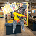 Ein als Wildschwein verkleideter Mensch sitzt auf einem Kassenband in einem Supermarkt. Er hält ein Schild mit der Aufschrift "Los für Vereine" und "Billa" in den Händen. Im Hintergrund sind Einkaufsregale zu sehen.