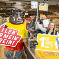Eine Frau in einem Supermarkt hält ein Schild mit der Aufschrift "1 Los für Vereine". Neben ihr steht eine Person in einem Wildschwein-Kostüm, die ein weiteres Schild mit der Aufschrift "I leb' für mein Verein!" hält. Im Hintergrund sind Regale mit Lebensmitteln zu sehen.