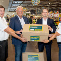 Eine Gruppe von vier Personen in einem Supermarkt hält Kartons mit der Aufschrift „Voll Versorgt“. Im Hintergrund sind Regale mit Obst und Gemüse zu sehen. Alle tragen formelle Kleidung oder Polo-Shirts.