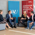 Eine Gruppe von vier Personen sitzt auf Sofas und unterhält sich. Im Hintergrund befinden sich zwei Werbebanner, eines mit dem Logo der WU Wien und das andere mit "Gemeinsam für ein besseres Leben". Die Personen sind formell gekleidet.