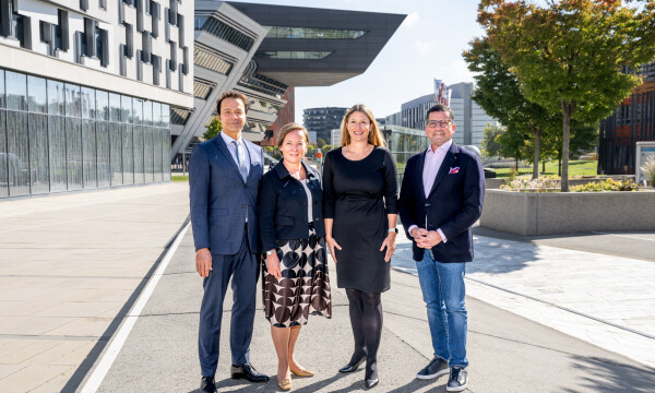 Vier Personen stehen nebeneinander auf einem Gehweg, mit modernen Gebäuden im Hintergrund. Drei tragen Bürokleidung, während eine Person leger gekleidet ist. Es ist ein sonniger Tag.
