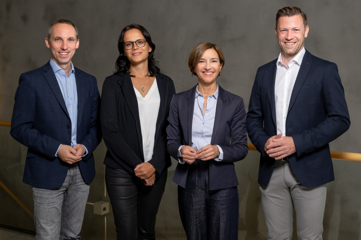 Vier Personen in Businesskleidung lächeln in die Kamera. Eine Frau trägt eine Brille und lässige Kleidung, die anderen sind formell gekleidet. Im Hintergrund ist eine graue Wand.