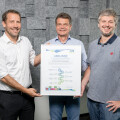 Drei Männer stehen lächelnd in einem Raum und halten gemeinsam eine Urkunde. Zwei von ihnen tragen Hemden, der dritte ein helles Polo-Shirt. Der Hintergrund besteht aus einer grauen Wand mit Akustikplatten.