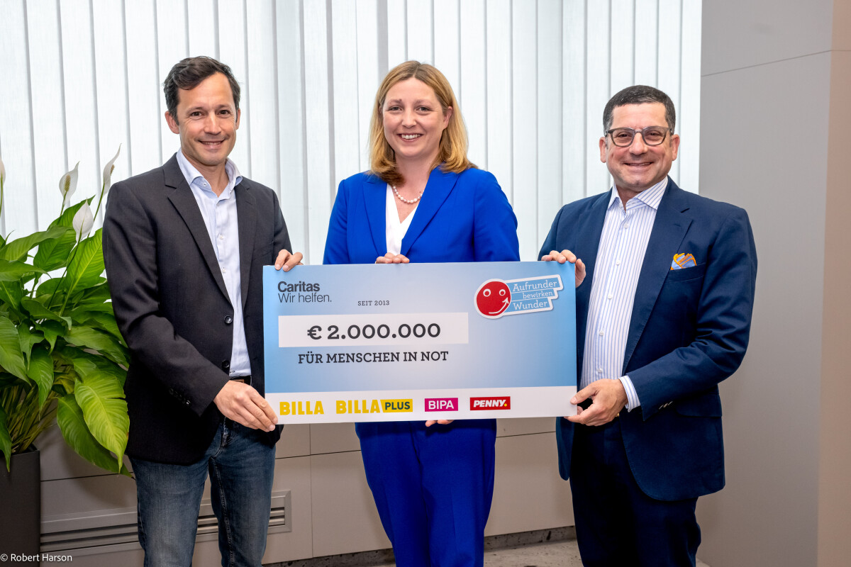 Drei Personen halten einen großen Scheck über 2.000.000 Euro für Menschen in Not. Links steht ein Mann in einem Blazer, in der Mitte eine Frau in einem blauen Blazer, und rechts steht ein weiterer Mann in einem Anzug. Im Hintergrund befinden sich Pflanzen.
