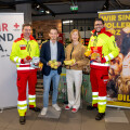 Eine Gruppe von vier Personen steht in einem Lebensmittelgeschäft. Zwei von ihnen tragen Rettungsdienst-Uniformen und halten Lebensmittelprodukte. Die anderen zwei sind in Freizeitkleidung und halten ebenfalls Produkte. Im Hintergrund sind Werbetafeln mit der Aufschrift "WIR SIND DA." und "WIR SIND VOLLER HERZ" sichtbar.
