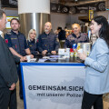 Polizistinnen und Polizisten stehen hinter einem Infostand einer Polizeikampagne in einem Geschäft. Ein Mann im Anzug und eine Frau im blauen Blazer sprechen mit ihnen. Der Tisch ist mit Werbematerialien bedeckt. Im Hintergrund sind Regale mit Lebensmitteln und Schilder zu sehen.