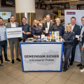 Mehrere Menschen stehen vor einem Tisch mit dem Banner "Gemeinsam.Sicher mit unserer Polizei". Einige tragen Polizeiuniformen, andere sind in Zivilkleidung. Im Hintergrund sind unter anderem ein Pizza- und ein Leberkäse-Bereich zu sehen.