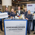 Personengruppe in einem Supermarkt vor einem Stand mit der Aufschrift "GEMEINSAM.SICHER mit unserer Polizei". Mehrere Menschen, darunter Polizeiangehörige in Uniform, lächeln in die Kamera und halten verschiedene Gegenstände. Im Hintergrund sind Ladenregale und eine Pizza-Anzeige zu sehen.