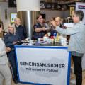 Eine Gruppe von Polizisten und Personen steht an einem Informationsstand in einem Einkaufszentrum. Der Stand trägt den Schriftzug "GEMEINSAM.SICHER mit unserer Polizei". Ein Mann spricht mit den Polizisten, während eine Frau Broschüren hält. Im Hintergrund sind Regale mit Produkten zu sehen.