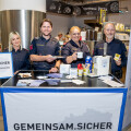 Vier Polizeibeamte stehen hinter einem Infostand im Supermarkt, lächeln und präsentieren Informationsmaterial und Tassen. Der Stand ist mit einem Banner "GEMEINSAM.SICHER" gekennzeichnet.