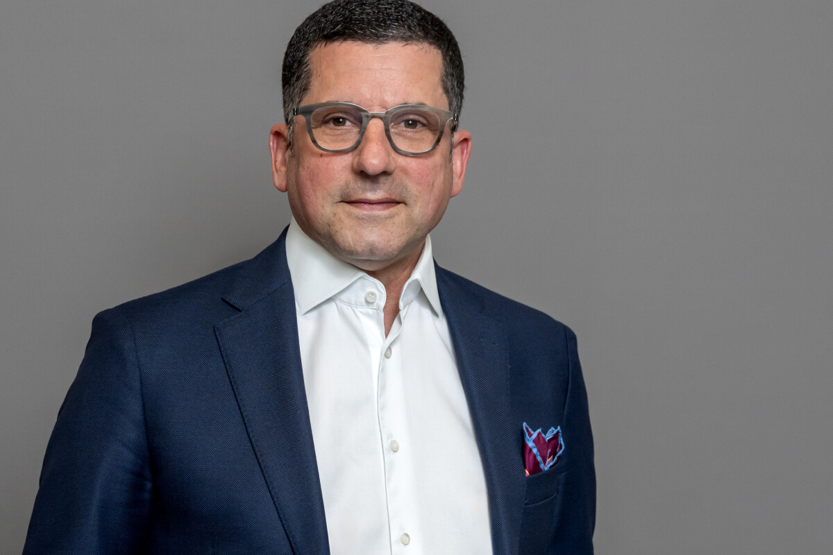 Ein Mann in einem dunklen Anzug und weißem Hemd steht vor einem grauen Hintergrund. Er trägt eine Brille und hat ein Einstecktuch in seiner Brusttasche.