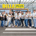 Eine Gruppe von Menschen steht vor einem BILLA PLUS Supermarkt. Sie tragen uniforme Kleidung und lächeln in Richtung Kamera. Einige Personen zeigen Gesten des Daumens hoch. Der Eingang des Supermarkts ist im Hintergrund zu sehen.