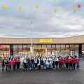 Eine Gruppe von Menschen steht vor einem großen Supermarktgebäude mit der Aufschrift "BILLA". Im Hintergrund sind rote und gelbe Luftballons am Himmel zu sehen. Der Himmel ist teils bewölkt.