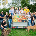 Gruppe von Menschen steht lächelnd um einen Anhänger mit gefüllten Säcken im Freien. Ein Hund steht vorne. Im Hintergrund sind Bäume und grünes Gras. Alle tragen lässige Kleidung und Sonnenbrillen oder Hüte.