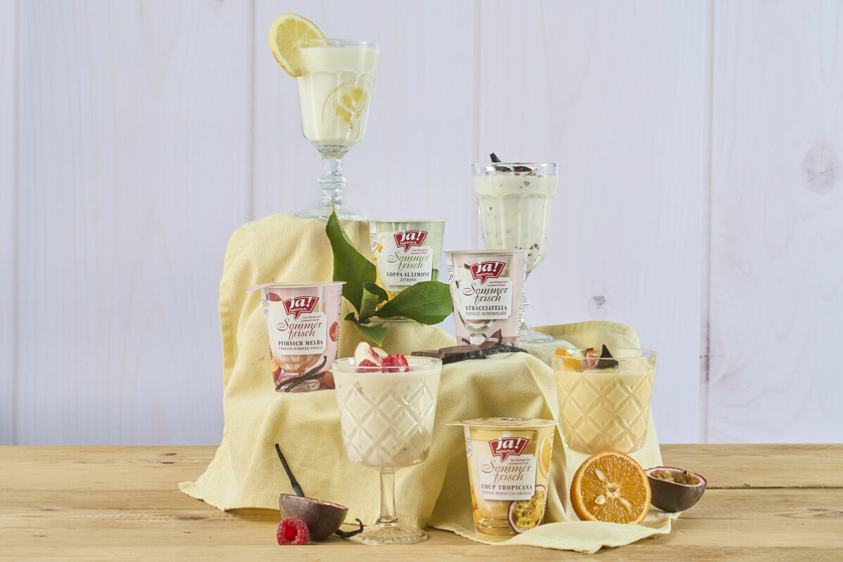 Eine Auswahl von Dessertgläsern, darunter Tiramisu und Panna Cotta, auf einem Tisch mit einem gelben Stoff. Dekorationen umfassen Zitronenscheiben, Passionsfrucht, Himbeeren und Schokoladenstückchen.