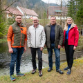 Vier Personen stehen vor einem Fluss in einer ländlichen Umgebung. Im Hintergrund sind Bäume und Gebäude zu sehen. Die Personen tragen Outdoor-Kleidung und lächeln in die Kamera.