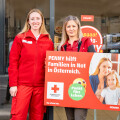 Zwei Frauen in roten Uniformen stehen vor einem Geschäft. Eine von ihnen hält ein Schild mit der Aufschrift "PENNY hilft Familien in Not in Österreich" und einem Logo des Roten Kreuzes.