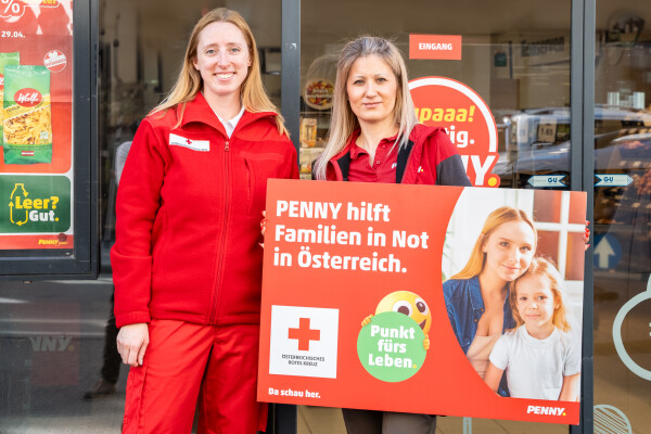 Zwei Frauen in roten Uniformen stehen vor einem Geschäft. Eine von ihnen hält ein Schild mit der Aufschrift "PENNY hilft Familien in Not in Österreich" und einem Logo des Roten Kreuzes.