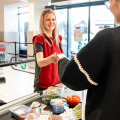 Eine Kassiererin in roter Uniform nimmt eine Karte von einem Kunden entgegen. Auf dem Kassenband liegen verschiedene Lebensmittel wie Gemüse, Obst und verpackte Waren. Im Hintergrund sind Einkaufswagen und ein Fenster mit Tageslicht zu sehen.