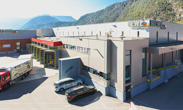 Großes Industriegebäude mit der Aufschrift "REWE" auf dem Dach, umgeben von Bergen. Ein roter LKW und zwei Autos sind auf dem Parkplatz zu sehen. Sonniges Wetter mit klarer Sicht.