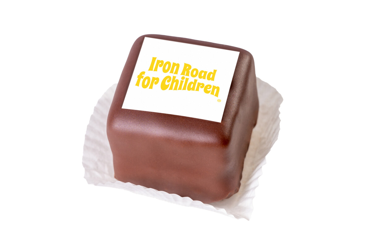 Ein Würfelkuchen mit Schokoladenüberzug, auf dem ein weißes Rechteck mit der Aufschrift "Iron Road for Children" in gelber Schrift liegt. Der Kuchen steht auf einem weißen Papier.