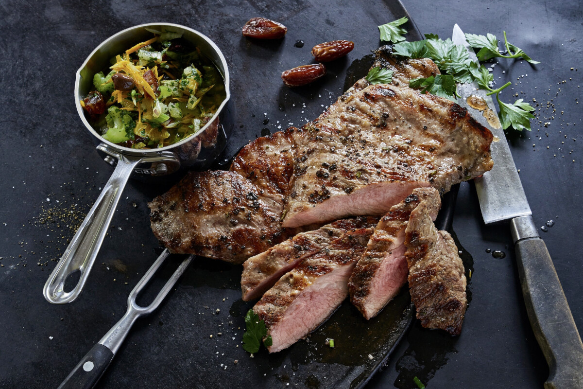 Gegrilltes Rindersteak, in Scheiben geschnitten, liegt auf einer dunklen Platte neben einem Messer. Daneben ein Topf mit Gemüsebeilage, darunter Brokkoli und Paprika, garniert mit Petersilie und Datteln.