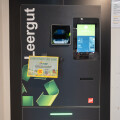 Leergut-Automat mit grünem Bildschirm und recyclebarem Logo. Ein Plakat mit der Aufschrift „Dein Pfand fürs Land“ ist angebracht.