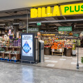 Eingang eines Billa Plus Supermarkts. Im Vordergrund stehen ein Weihnachtsbaum und ein Werbeaufsteller. Im Inneren sieht man Obst, Gemüse und andere Lebensmittel.