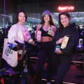 Drei lächelnde Frauen stehen neben Trainingsrädern in einem Fitnessstudio, halten Getränkepackungen in der Hand. Neonlicht im Hintergrund.