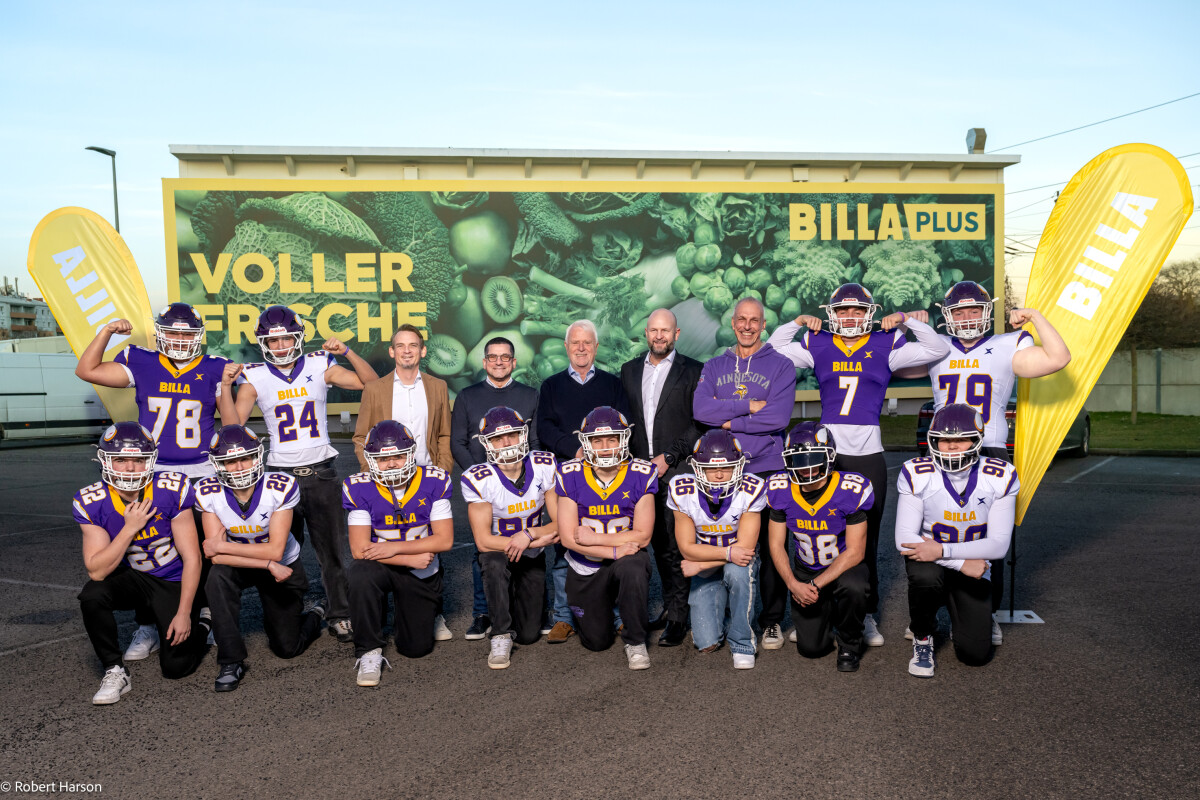 Eine Gruppe von American-Football-Spielern in lila-weißen Trikots posiert mit drei Männern in Anzügen vor einer BILLA-Werbetafel. Sie stehen auf einem Parkplatz, flankiert von gelben BILLA-Flaggen.