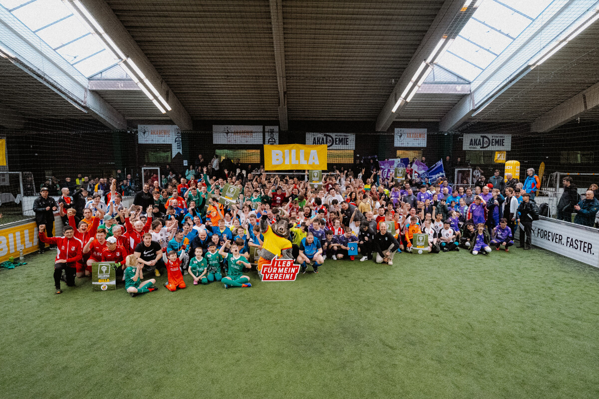 Gruppe von Kindern und Erwachsenen in bunten Sporttrikots posiert auf einem Indoor-Spielfeld. Sie halten Schilder und Trophäen. Im Hintergrund sind Werbeplakate zu sehen.