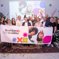 Eine Gruppe von Menschen hält ein Banner mit der Aufschrift "NextGen Buddies" in einem hellen Raum. Sie feiern gemeinsam und stehen auf Konfetti. Im Hintergrund sind Pflanzen und eine Projektion an der Wand zu sehen. Alle wirken fröhlich.