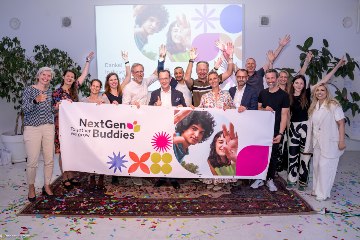 Eine Gruppe von Menschen hält ein Banner mit der Aufschrift "NextGen Buddies" in einem hellen Raum. Sie feiern gemeinsam und stehen auf Konfetti. Im Hintergrund sind Pflanzen und eine Projektion an der Wand zu sehen. Alle wirken fröhlich.