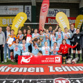 Eine Damenfußballmannschaft feiert den Sieg bei einem Turnier. Die Spielerinnen tragen Medaillen und halten eine Trophäe. Sie stehen auf einem Indoor-Spielfeld vor mehreren Werbebannern und posieren fröhlich für das Foto. Rechteckige Schilder mit Sponsorenlogos sind sichtbar.