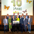 Eine Gruppe von Menschen steht zusammen und lächelt vor einem Hintergrund mit der Aufschrift "10 Jahre" und Schmetterlingsabbildungen. Unten halten zwei Personen ein Schild mit dem Logo von "Billa Blühendes Österreich." Die Gruppe ist formell gekleidet.