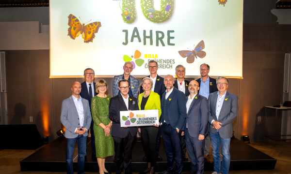 Eine Gruppe von Menschen steht zusammen und lächelt vor einem Hintergrund mit der Aufschrift "10 Jahre" und Schmetterlingsabbildungen. Unten halten zwei Personen ein Schild mit dem Logo von "Billa Blühendes Österreich." Die Gruppe ist formell gekleidet.