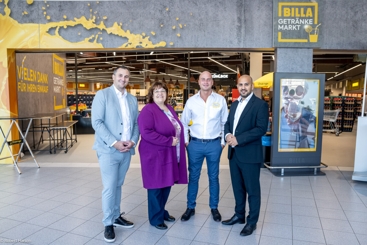 Vier Personen stehen vor einem BILLA Getränkemarkt. Zwei Männer tragen Anzüge, eine Frau trägt eine lila Jacke, und ein Mann trägt ein weißes Hemd mit einem BILLA-Logo. Im Hintergrund sind Einkaufswagen und Regale zu sehen.