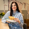 Eine Frau mit langen, dunklen Haaren hält ein Croissant auf einem Teller und lächelt. Sie trägt eine Jeansjacke und sitzt an einem Tisch mit einer Pflanze im Hintergrund. Die Wand hat ein abstraktes, gemustertes Design.