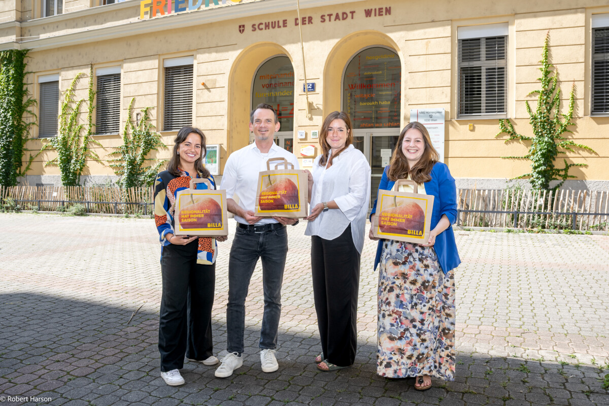 Vier Personen stehen vor einem Schulgebäude und halten Papiertüten mit dem Logo „Goldenes Wiener Herz“ darauf. Sie lächeln in die Kamera. Im Hintergrund ist die Fassade der Schule mit Fenstern und Efeu zu sehen.