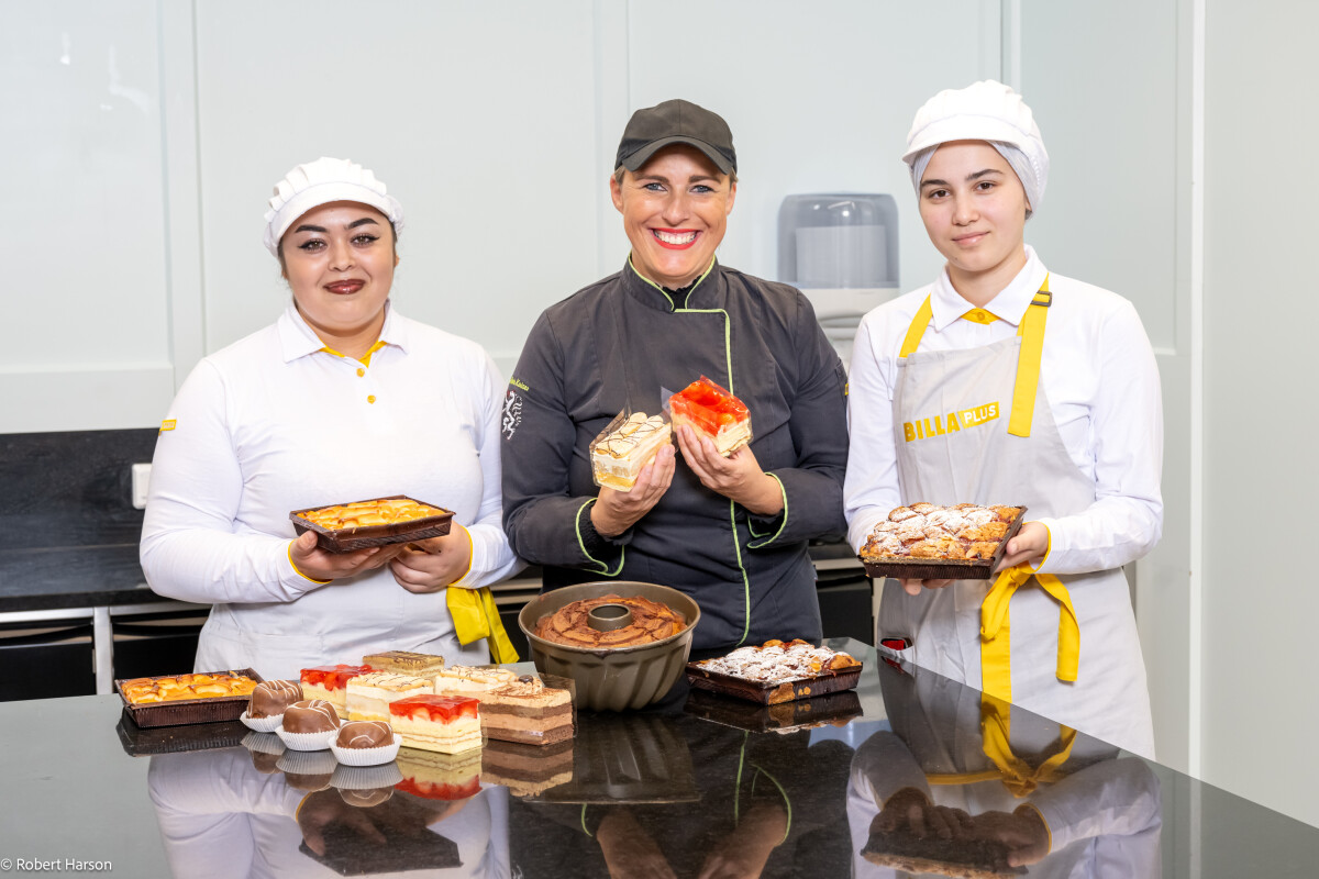 Drei Bäckerinnen in Uniformen stehen in einer Küche und präsentieren stolz ihre gebackenen Kuchen und Desserts. Sie tragen weiße Kappen und Schürzen, ein paar im Bild hat schwarze Details. Vor ihnen stehen verschiedene Kuchen auf einem Tisch.