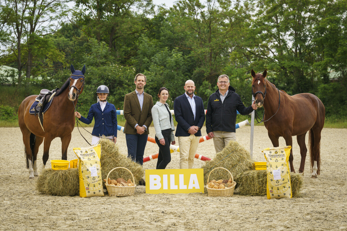 Eine Gruppe von fünf Menschen steht auf einem Reitplatz zwischen zwei Pferden. Vor ihnen liegen Heuballen und Körbe mit Kartoffeln, daneben steht ein gelbes Schild mit der Aufschrift "BILLA". Im Hintergrund sind Bäume sichtbar.