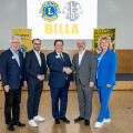 Fünf Personen stehen lächelnd in einem Konferenzraum. Zwei von ihnen schütteln sich die Hände. Im Hintergrund sind Logos von Lions und Leo Clubs sowie der Schriftzug "BILLA" zu sehen. Alle tragen formelle Kleidung.