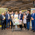 Eine Gruppe von Menschen steht um einen Einkaufswagen in einem Supermarkt. Der Wagen ist mit verschiedenen Lebensmittelprodukten gefüllt. Im Hintergrund sind Banner mit Texten zu sehen. Alle Personen tragen formelle Kleidung und lächeln in die Kamera.