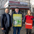 Drei Personen stehen vor einem Gebäude mit Herbstlaub auf dem Boden. Die zentrale Person hält ein Schild mit der Aufschrift "Hier gibt’s Pfand zurück" und Flaschenabbildungen. Die Person rechts hält ein Schild mit dem Logo von "Penny". Links steht eine weitere Person, die lächelt. Im Hintergrund ist das Schild eines Lokals mit dem Namen "Zum Scharfen Rene" sichtbar.